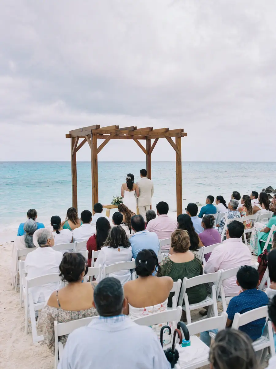 Invitados sentados en sillas blancas durante una boda en la playa, la pareja de pie bajo un arco de madera frente al mar